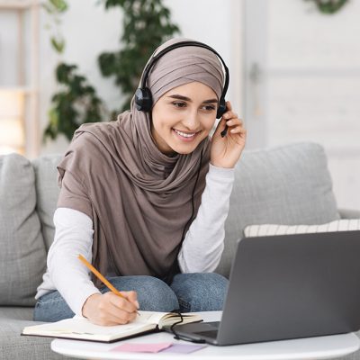 Online Education. Smiling Arabic Girl In Hijab Watching Webinar On Laptop At Home And Taking Notes, Using Headset